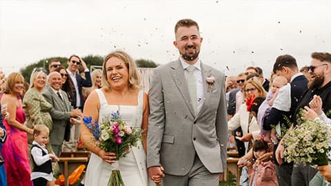A close up of Sian in her white wedding dress and Danny in his grey suit walking down the aisle in between friends and family who are all smiling.